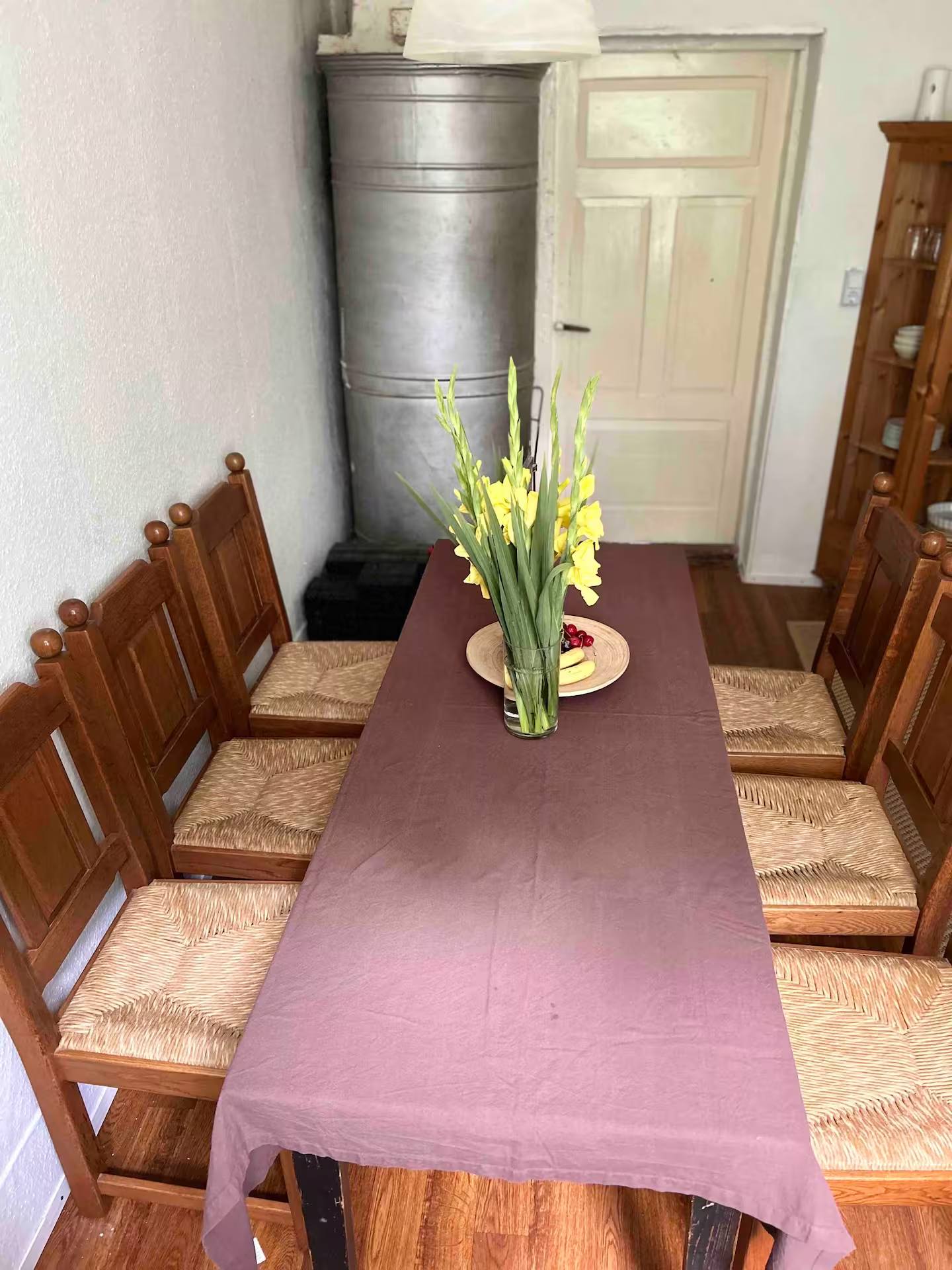Gedeckter Esstisch mit einer Vase gelber Gladiolen und einer Schale Obst, dahinter Holzstühle und ein Buffetschrank.