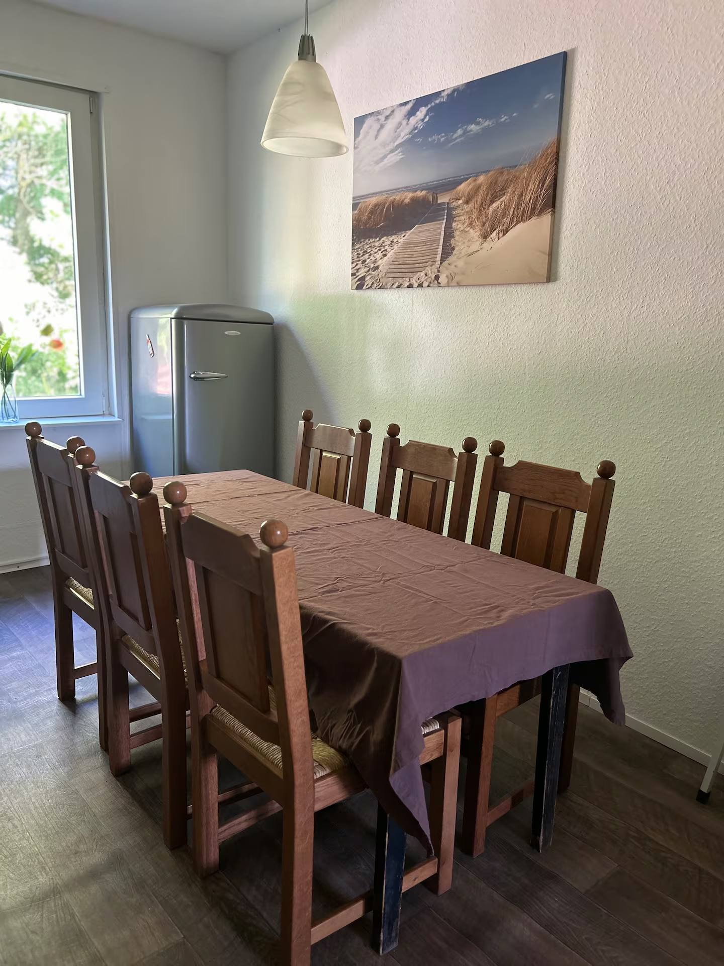 Esszimmer mit langem Holztisch, sechs hohen Holzstühlen und Tischdecke; darüber eine Pendelleuchte, daneben ein Kühlschrank im Retro-Design und ein Bild mit Dünenmotiv an der Wand.