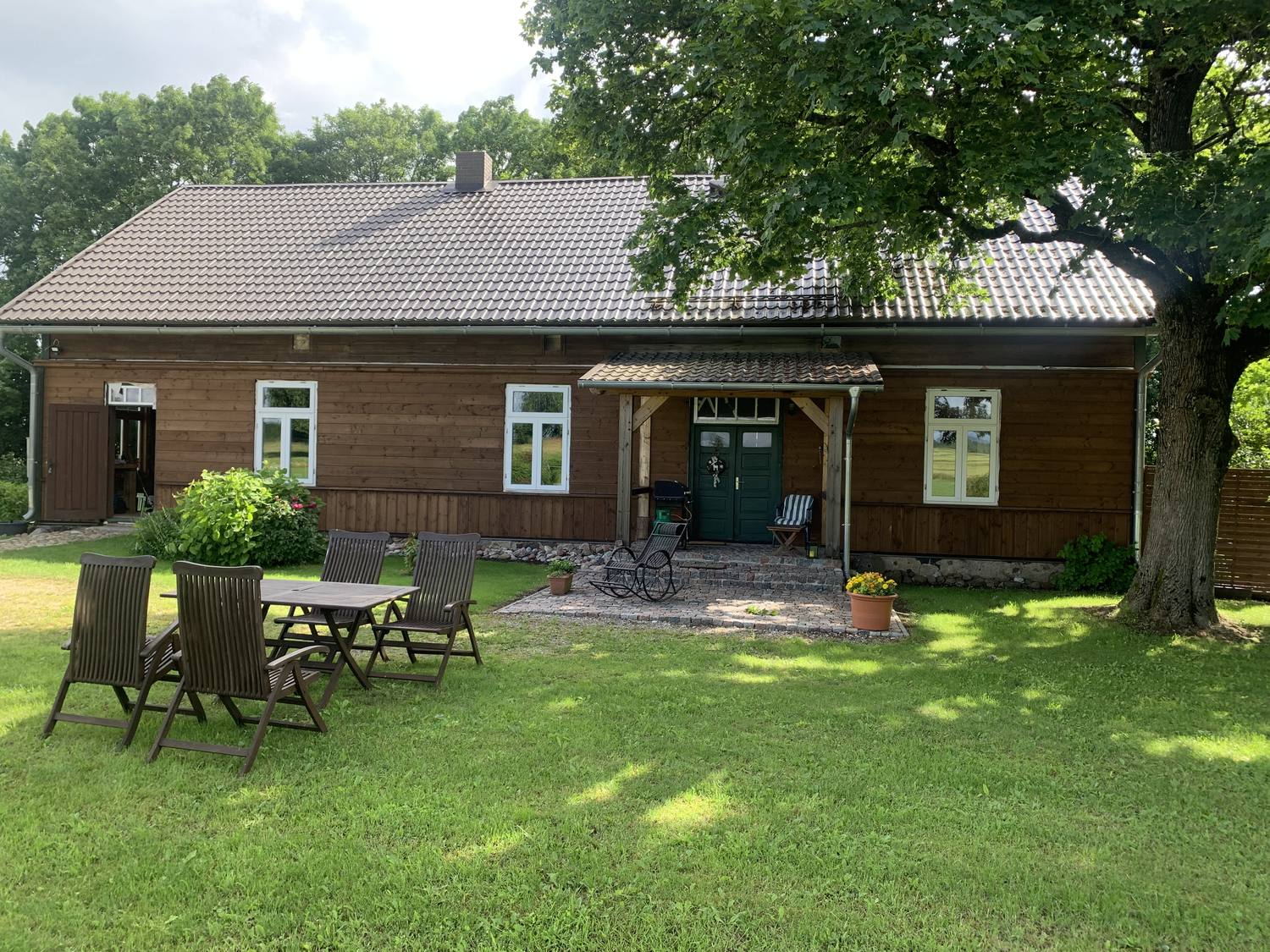 Langgestrecktes Holzhaus mit dunkel lasierter Bretterschalung, grauem Ziegeldach und überdachtem Eingang; davor ein Gartentisch mit Klappstühlen auf dem Rasen, rechts eine große Eiche.