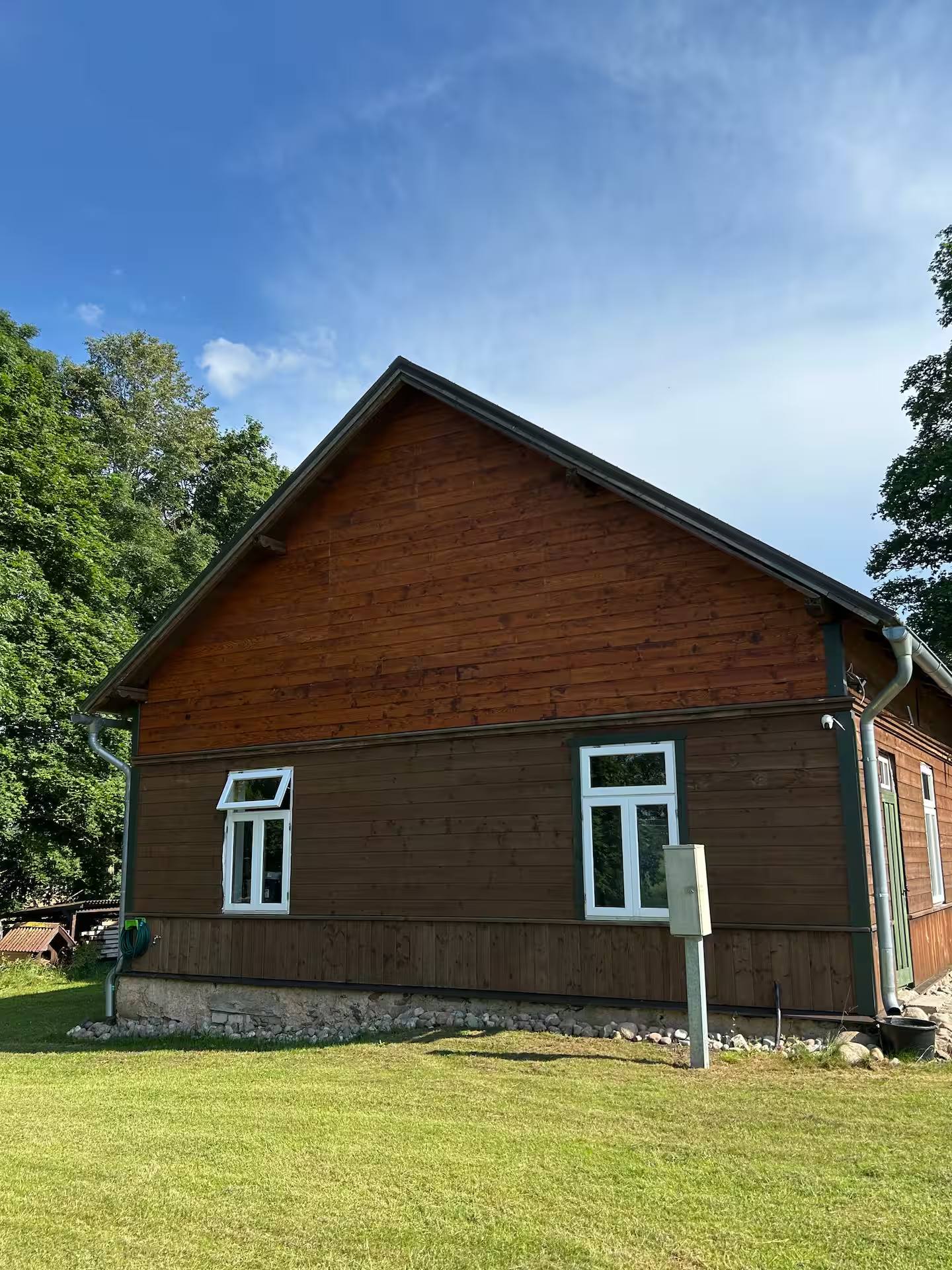 Giebelseite des Holzhauses mit zwei Fenstern, davor kurz gemähter Rasen und Bäume.