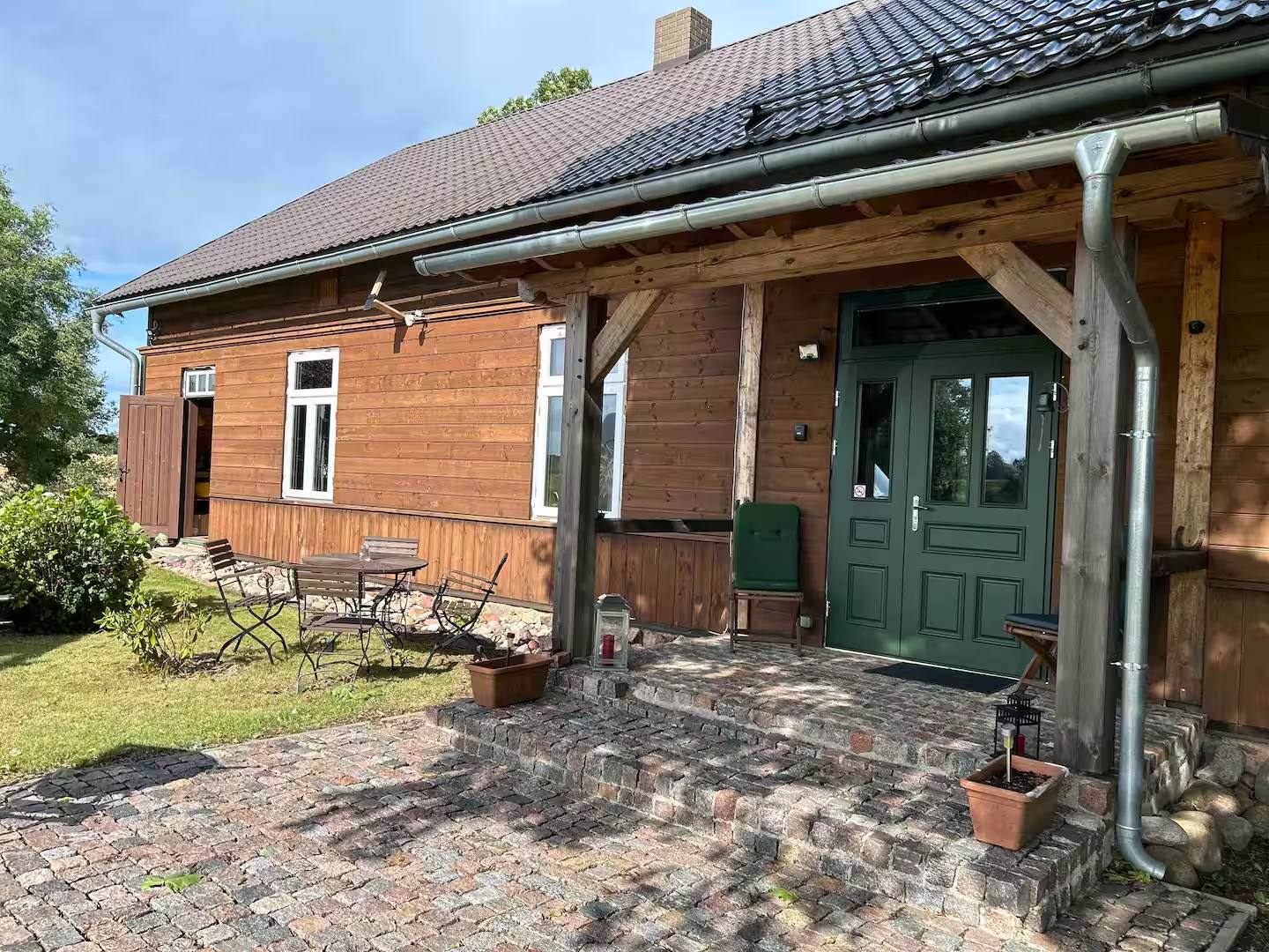 Längsseite des Hauses mit überdachtem Eingang, Gartenstühlen unter dem Vordach und Rasen bis an die Hauswand.