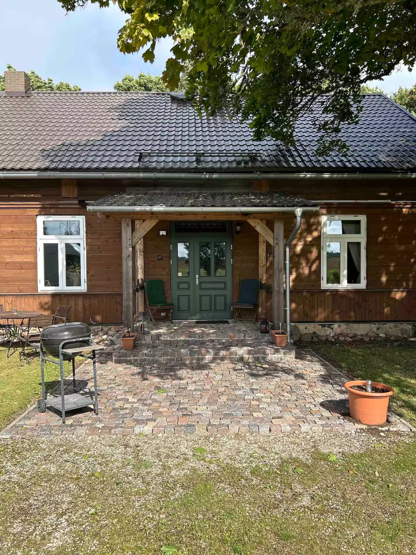Überdachter Hauseingang auf Holzstützen mit grüner Haustür; davor eine gepflasterte Fläche mit Kugelgrill und Blumentöpfen.