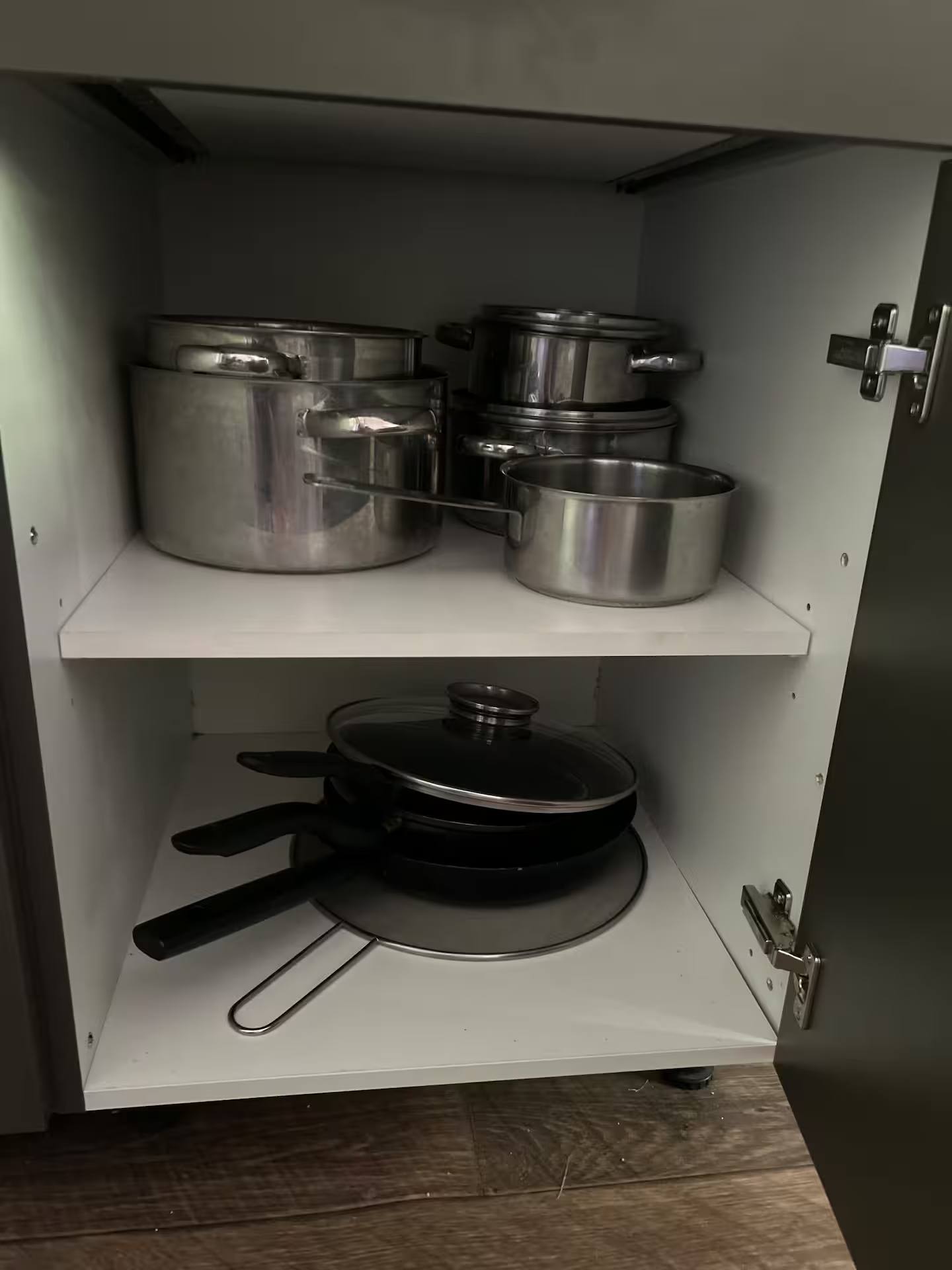 Geöffneter Küchenschrank mit gestapelten Edelstahltöpfen auf dem oberen und beschichteten Pfannen auf dem unteren Einlegeboden.