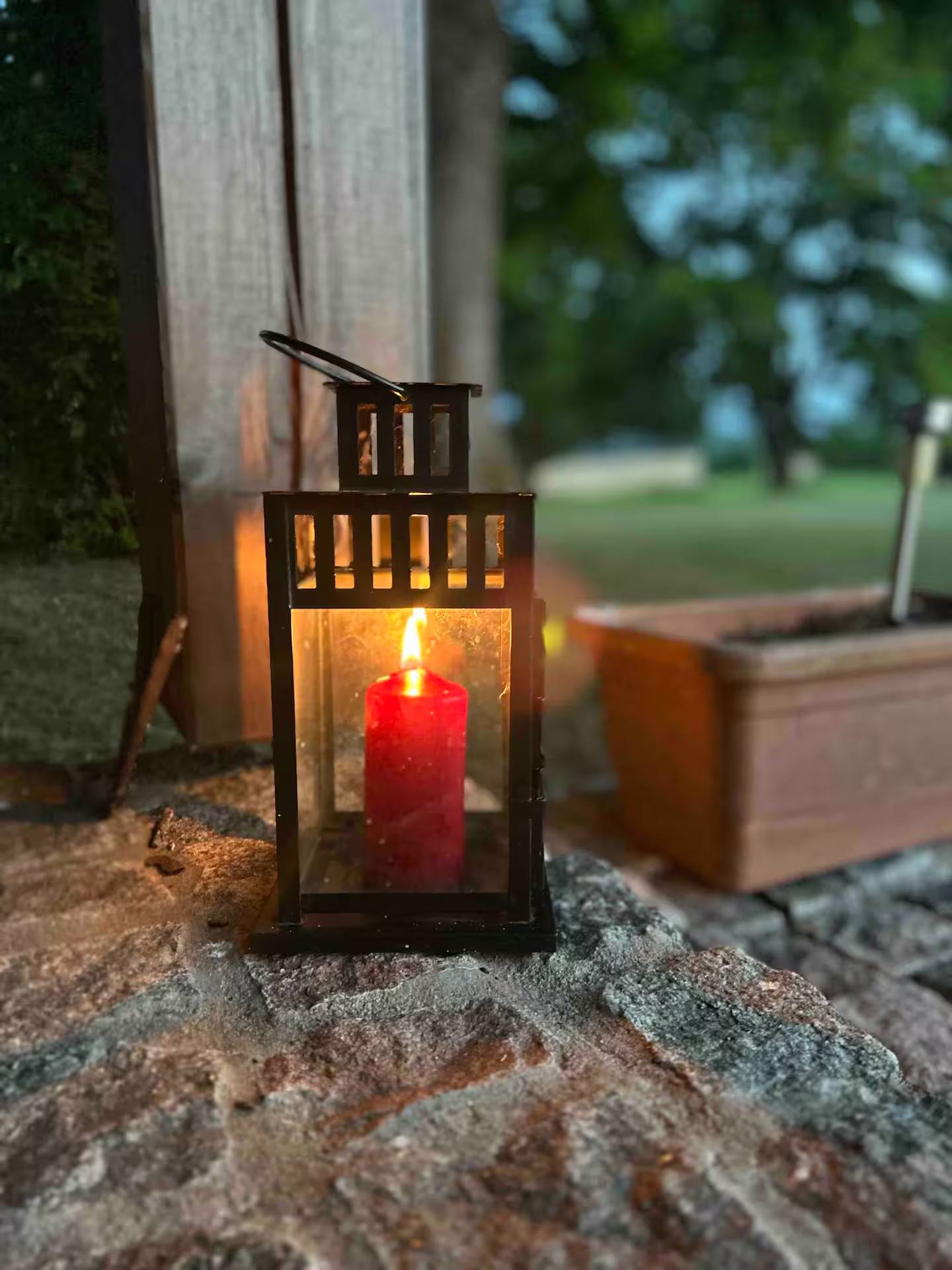 Metalllaterne mit brennender roter Kerze auf einer Steinplatte, daneben ein Holzblumenkasten, im Hintergrund der Rasen.