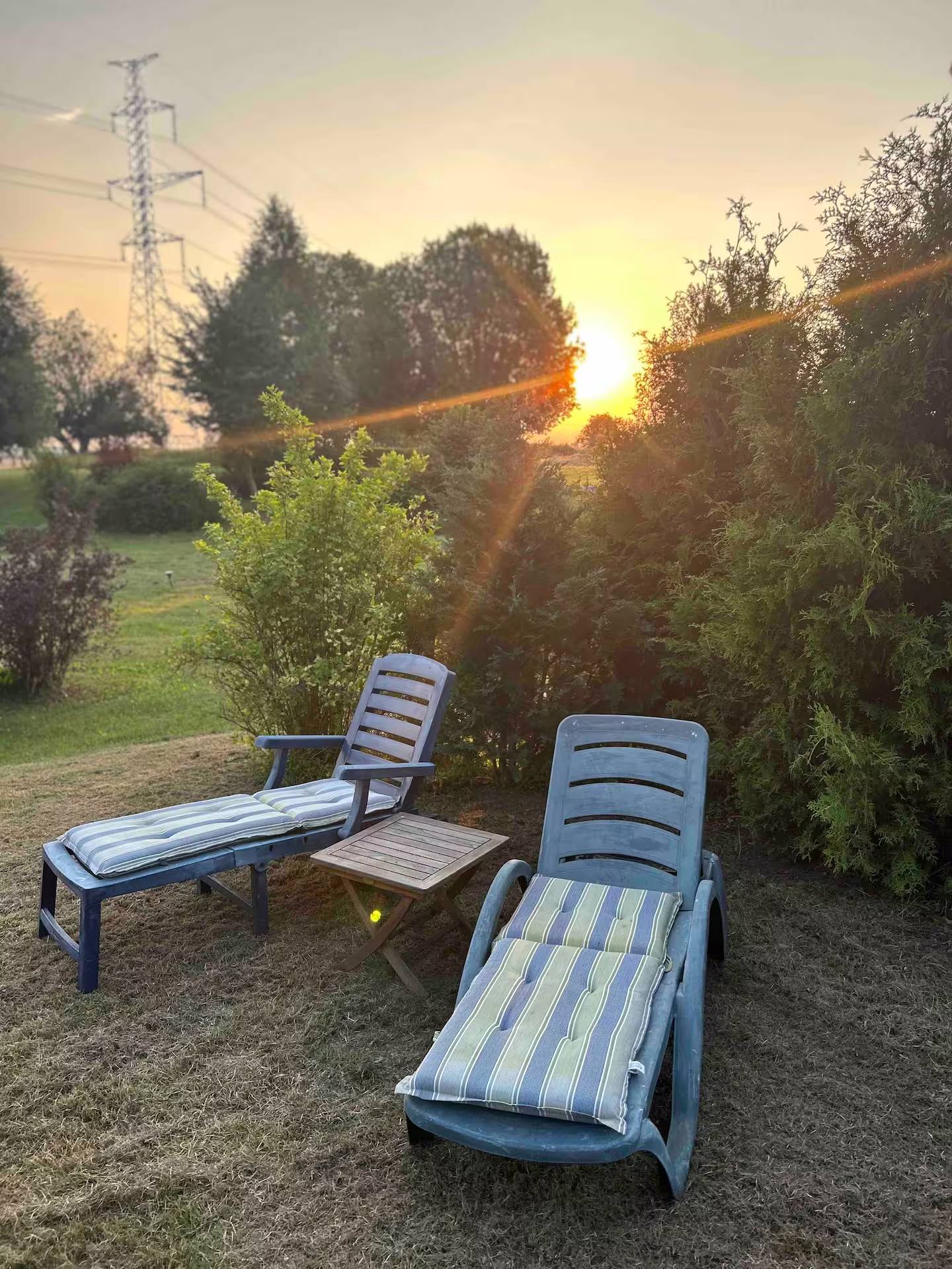 Zwei Sonnenliegen mit blau gestreiften Polstern auf dem Rasen, dahinter die tief stehende Abendsonne zwischen Bäumen.