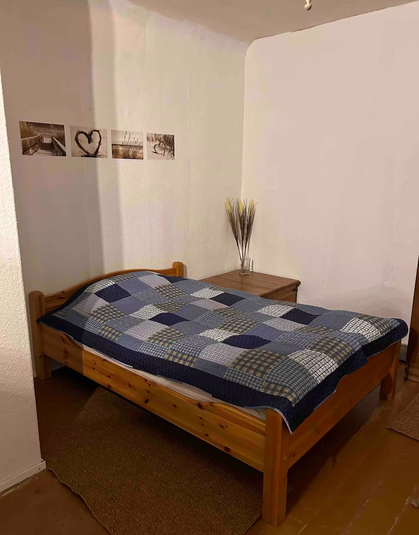 Schlafraum mit hellen Wänden: Holzbett mit blau-weiß kariertem Bezug, ein kleiner Nachttisch, drei gerahmte Fotos an der Wand.