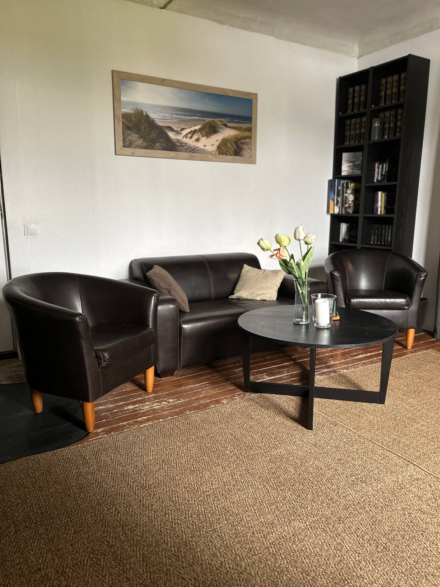 Wohnzimmer mit Ledersofa und zwei Ledersesseln um einen runden Couchtisch, Sisalteppich auf Holzdielen, ein Regal mit Büchern und ein Bild mit Dünenmotiv an der Wand.